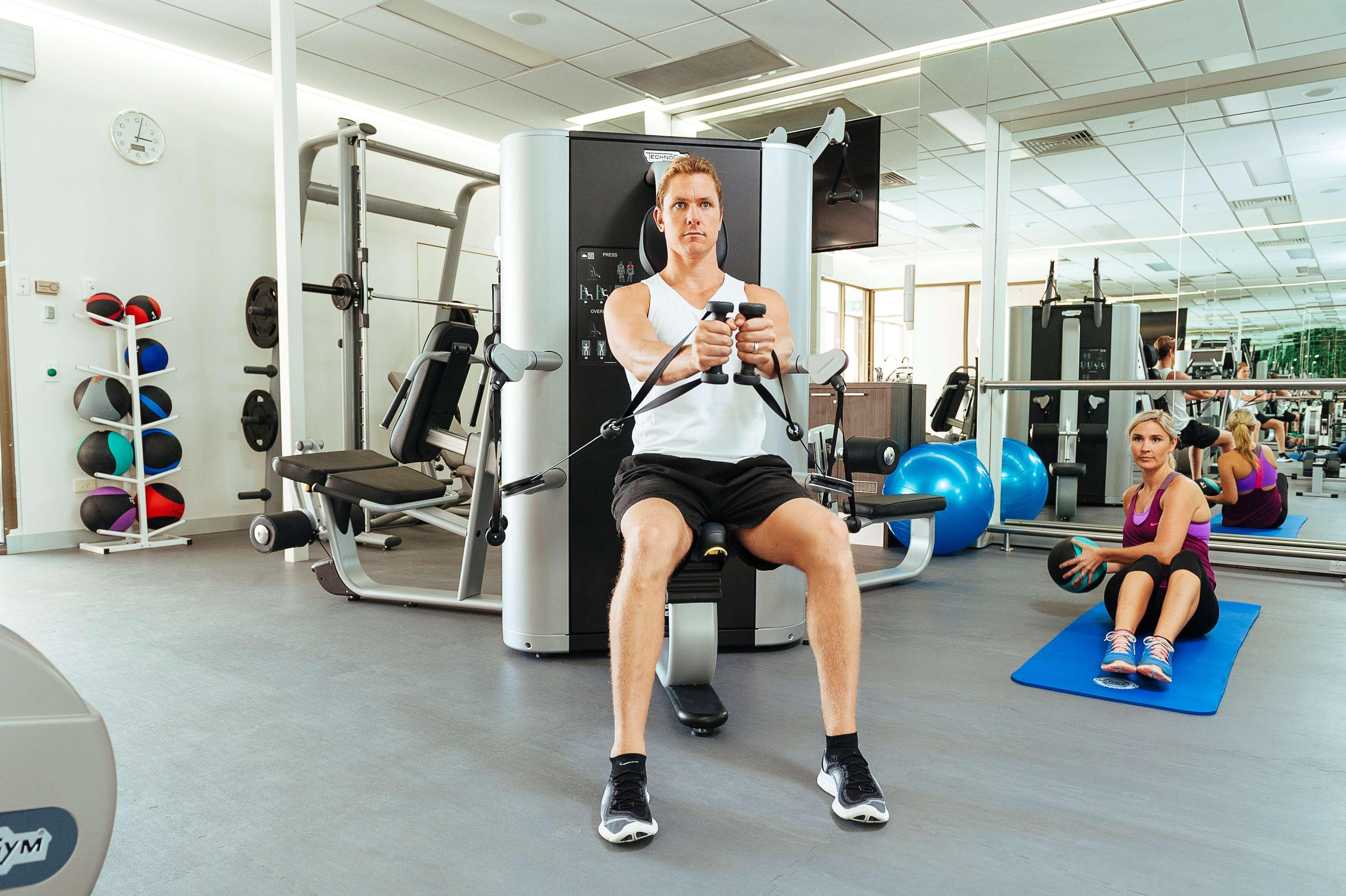 斯坦福德阿德莱德广场酒店 外观 照片 The photo shows a gym setting with two individuals engaged in workout activities. 

In the foreground, a man is seated on a machine, using the handles to perform a rowing exercise. He is wearing a sleeveless shirt and shorts, indicating an active wor