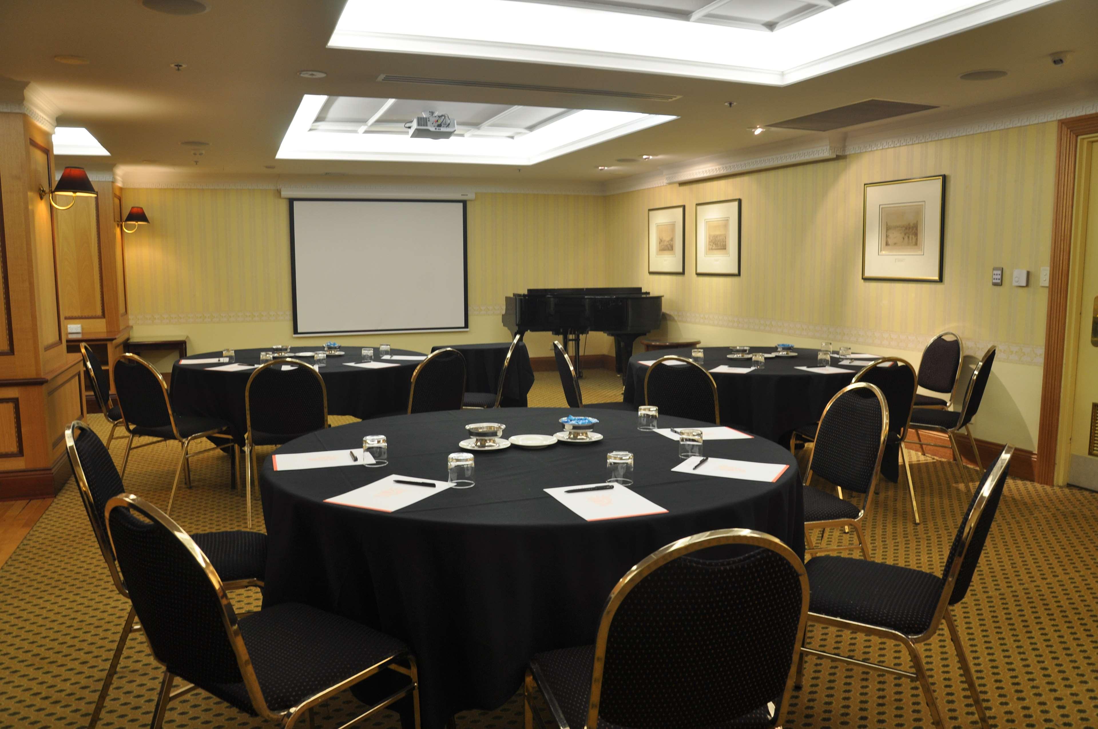 斯坦福德阿德莱德广场酒店 外观 照片 The photo shows a conference room set up for a meeting or presentation. There are several round tables covered with black tablecloths, each surrounded by black upholstered chairs. On the tables, there are notepads and glasses of water, indicating tha