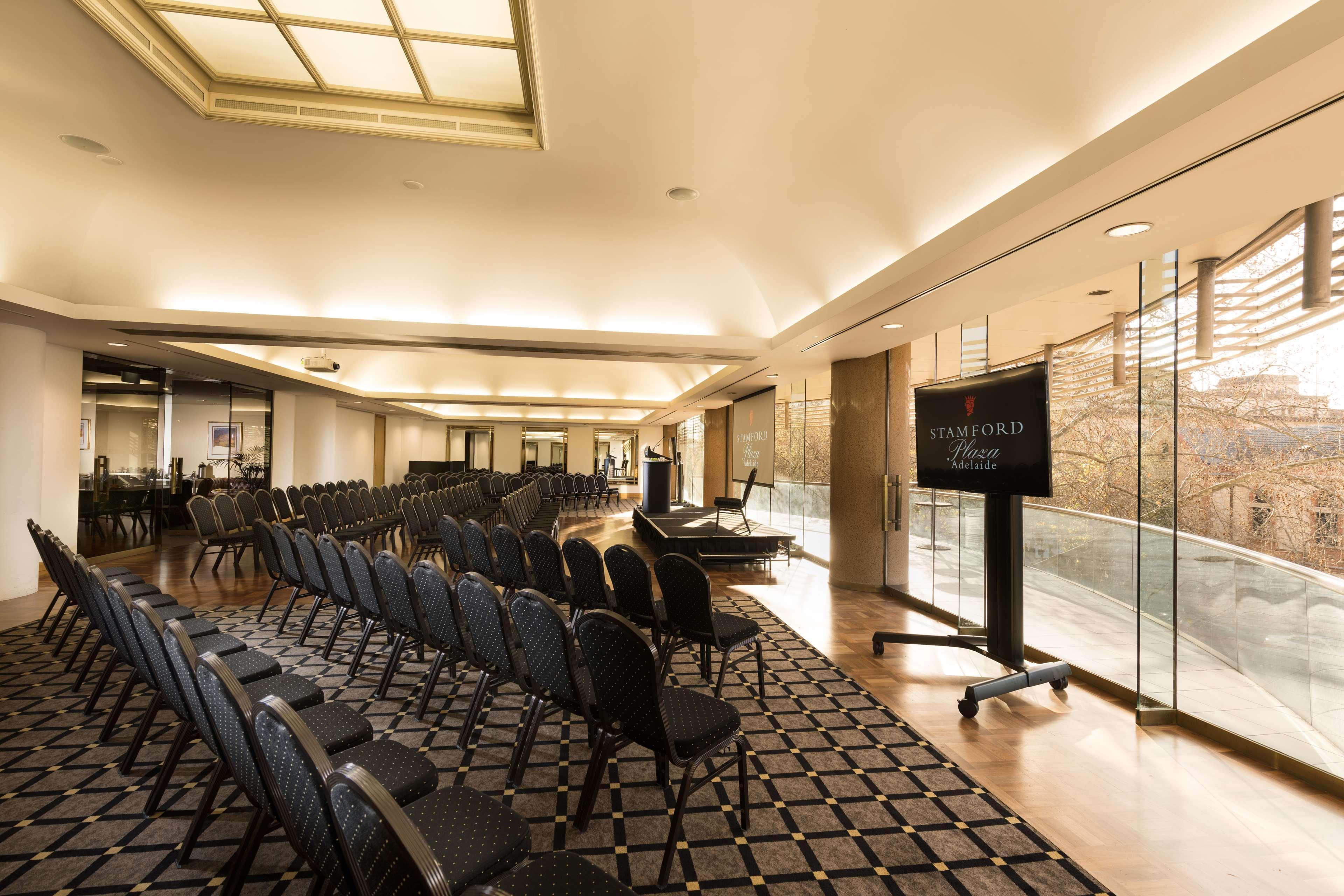 斯坦福德阿德莱德广场酒店 外观 照片 The photo shows a large, elegantly designed event space or meeting room. There are rows of black chairs set up facing a presentation area. The flooring features a patterned carpet, and large windows allow natural light to enter the room. On one side,