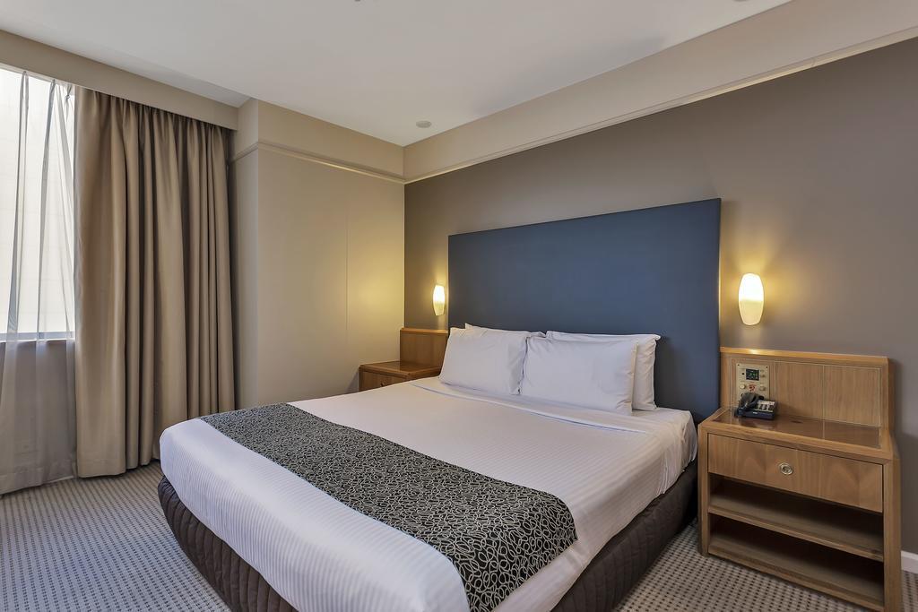 斯坦福德阿德莱德广场酒店 外观 照片 The photo shows a modern hotel room. It features a double bed with a white duvet and a decorative grey and black throw. The headboard is a solid dark color. There are two bedside tables on either side of the bed, each with a lamp. Natural light enter